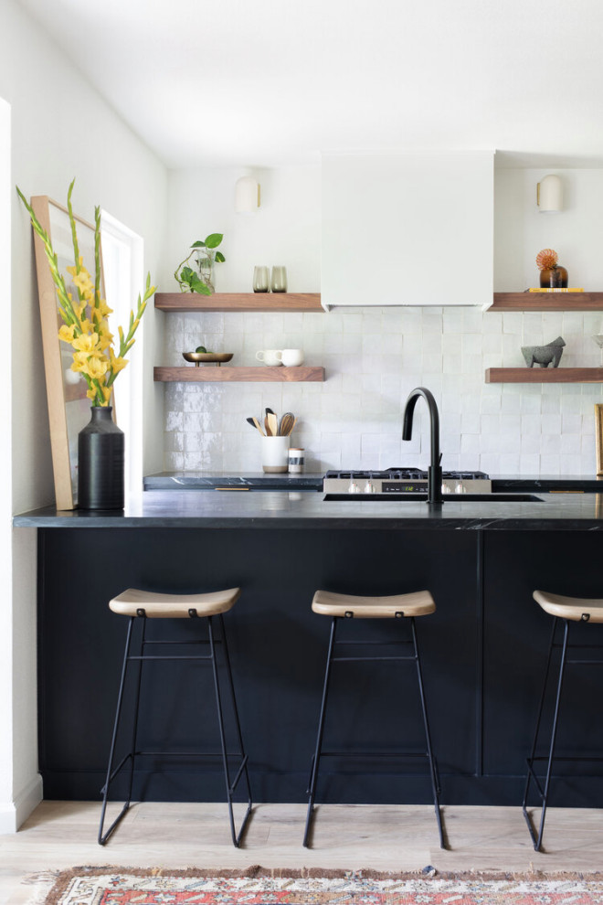 Example of a mid-sized danish galley light wood floor and beige floor open concept kitchen design in Austin with an undermount sink, flat-panel cabinets, black cabinets, soapstone countertops, white backsplash, ceramic backsplash, stainless steel appliances, an island and black countertops