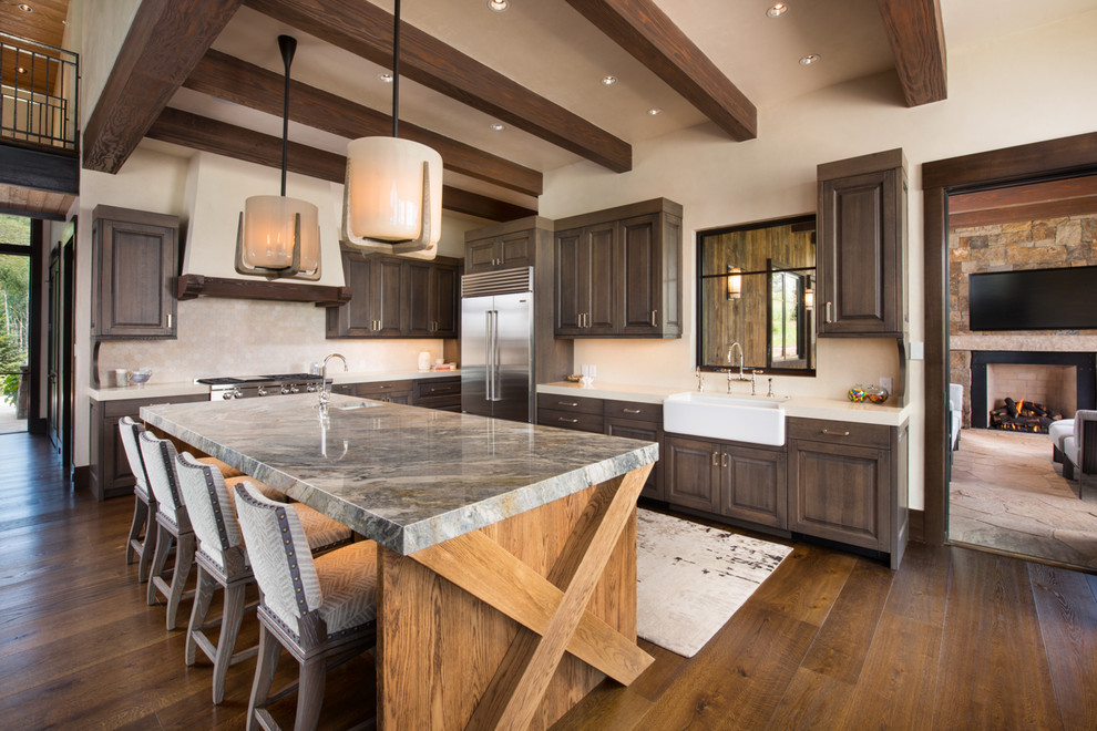 This is an example of an expansive country l-shaped kitchen in Denver with a farmhouse sink, raised-panel cabinets, quartzite benchtops, beige splashback, stone tile splashback, stainless steel appliances, with island, brown floor, dark wood cabinets and dark hardwood floors.