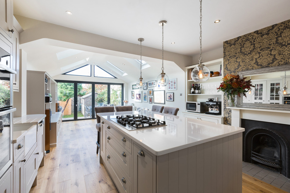 This is an example of a mid-sized contemporary single-wall open plan kitchen in London with a farmhouse sink, shaker cabinets, black appliances, with island, grey cabinets, light hardwood floors and beige floor.