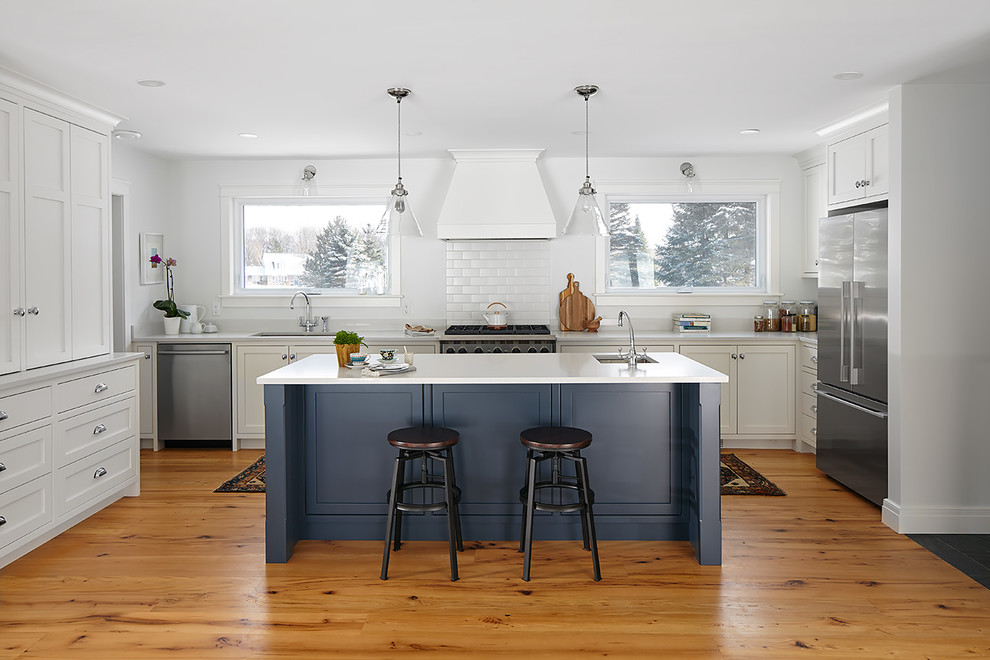 Example of a transitional medium tone wood floor kitchen design with an undermount sink, shaker cabinets, white cabinets, white backsplash, subway tile backsplash, stainless steel appliances, an island and white countertops