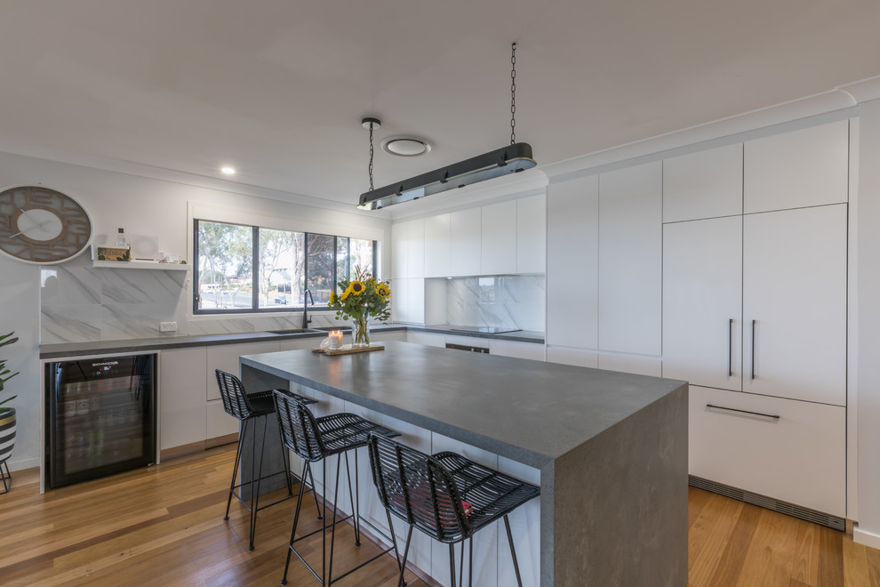 This is an example of a contemporary l-shaped kitchen in Sydney with an undermount sink, flat-panel cabinets, white cabinets, white splashback, stainless steel appliances, medium hardwood floors, with island, brown floor and grey benchtop.