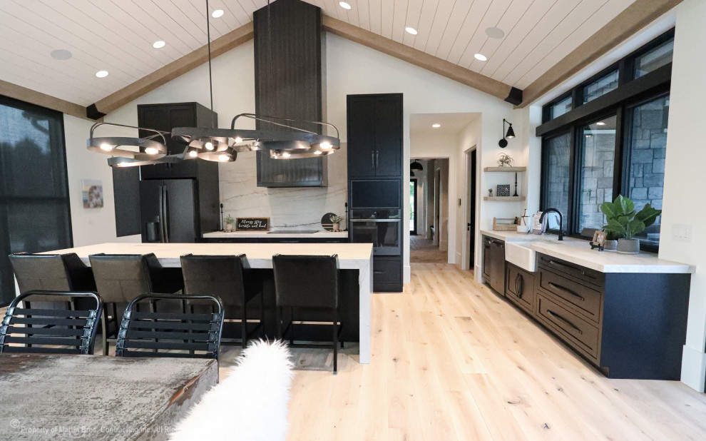 This is an example of a large industrial galley eat-in kitchen in Other with a farmhouse sink, shaker cabinets, black cabinets, quartzite benchtops, white splashback, stone slab splashback, black appliances, light hardwood floors, with island, white benchtop and timber.