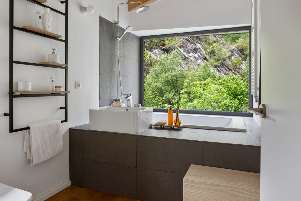 Large minimalist gray tile and stone slab medium tone wood floor, brown floor and exposed beam bathroom photo in Barcelona with gray cabinets, a one-piece toilet, white walls, a trough sink, tile countertops, gray countertops and a built-in vanity