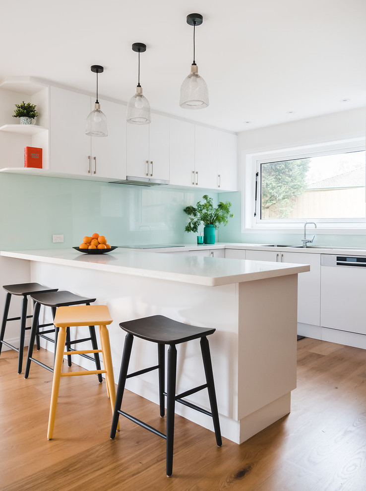 This is an example of a large contemporary u-shaped open plan kitchen in Melbourne with a double-bowl sink, flat-panel cabinets, white cabinets, granite benchtops, green splashback, glass sheet splashback, brown floor, white benchtop, panelled appliances, medium hardwood floors and a peninsula.