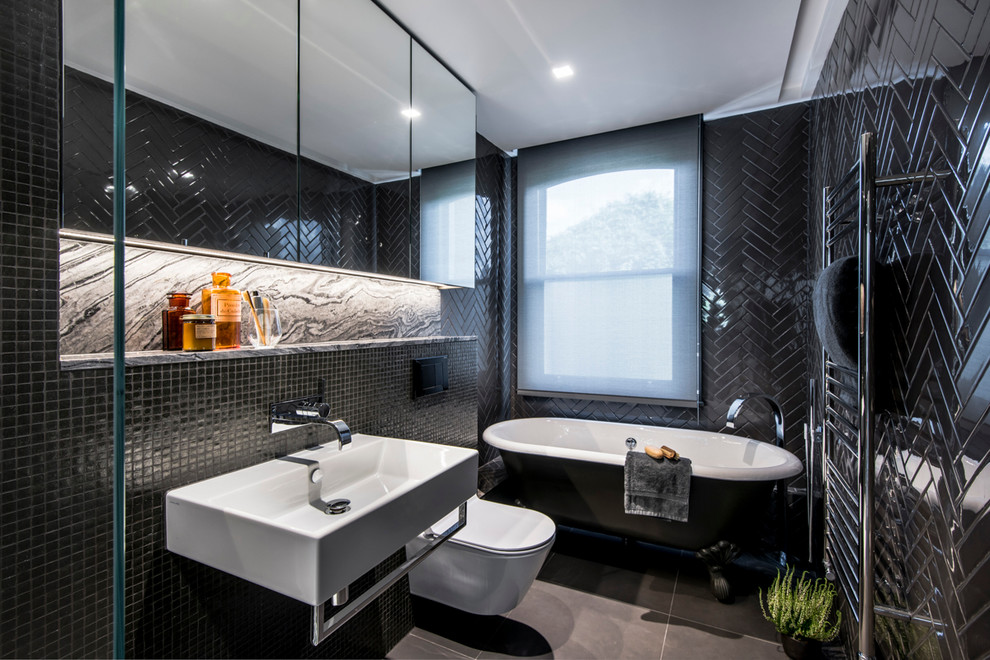 Photo of a contemporary bathroom in London with a claw-foot tub, a wall-mount toilet, black tile, black walls and a wall-mount sink.