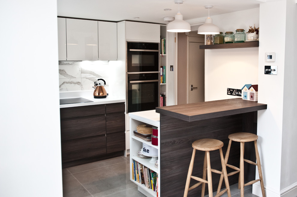 Large contemporary single-wall open plan kitchen in London with a drop-in sink, flat-panel cabinets, white cabinets, quartz benchtops, white splashback, ceramic splashback, black appliances, ceramic floors and a peninsula.