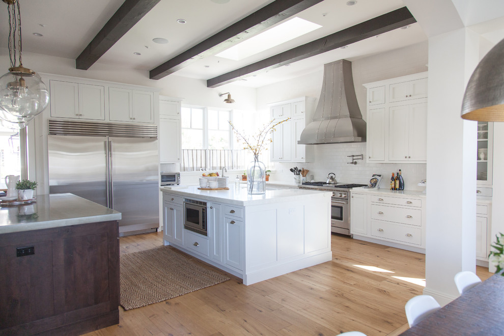Inspiration for a large transitional l-shaped light wood floor eat-in kitchen remodel in Phoenix with a farmhouse sink, white cabinets, white backsplash, stainless steel appliances, an island, stainless steel countertops, shaker cabinets and porcelain backsplash