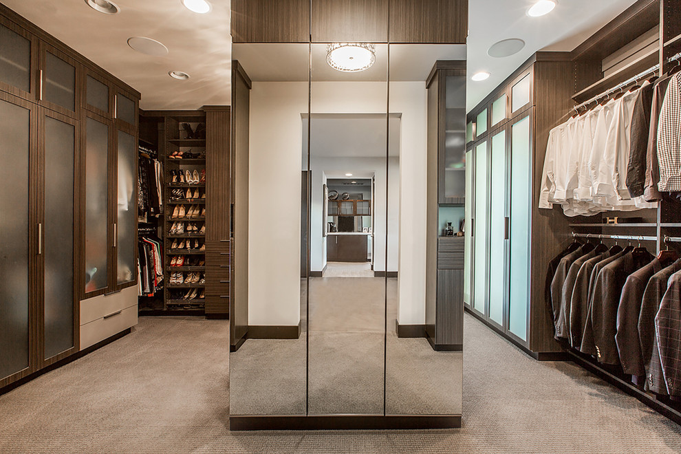 Expansive contemporary gender-neutral walk-in wardrobe in Las Vegas with glass-front cabinets, dark wood cabinets, carpet and brown floor.