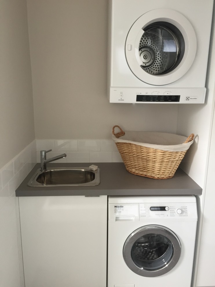 This is an example of a small contemporary single-wall utility room in Melbourne with a drop-in sink, flat-panel cabinets, white cabinets, quartz benchtops, grey walls, ceramic floors and a stacked washer and dryer.