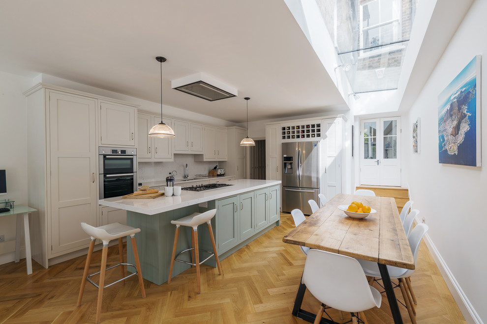 Mid-sized transitional l-shaped eat-in kitchen in London with light hardwood floors, with island, beige floor, an undermount sink, shaker cabinets, blue cabinets, white splashback and stainless steel appliances.