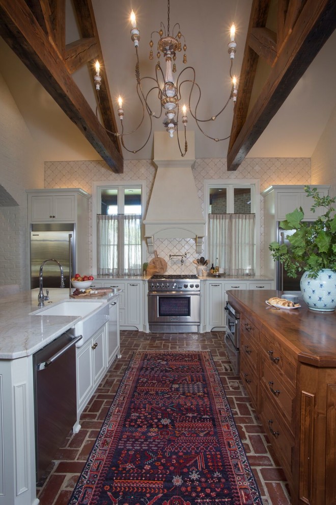 Mid-sized traditional galley separate kitchen in New Orleans with a farmhouse sink, recessed-panel cabinets, white cabinets, marble benchtops, white splashback, porcelain splashback, stainless steel appliances, brick floors and brown floor.