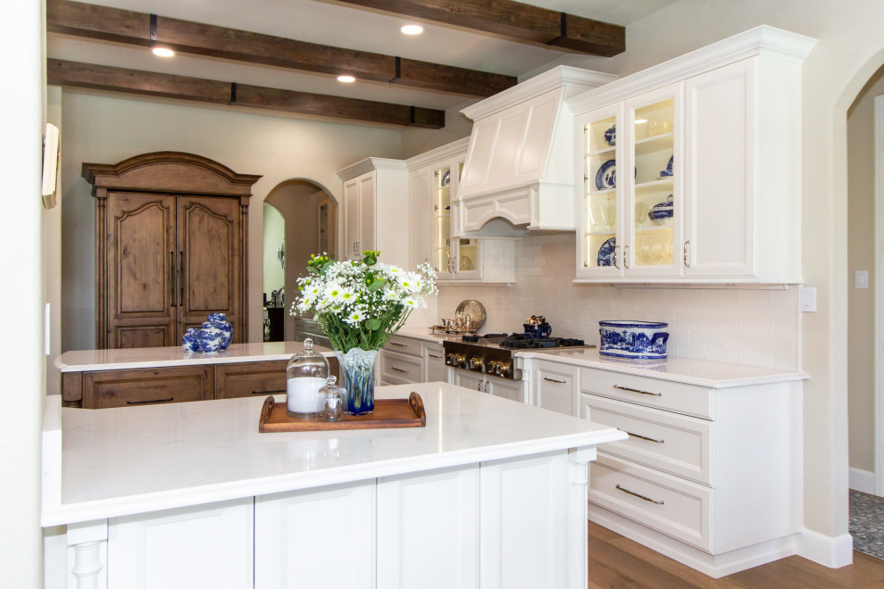 Inspiration for a large french country medium tone wood floor, brown floor and exposed beam kitchen remodel in Houston with an undermount sink, recessed-panel cabinets, white cabinets, quartz countertops, white backsplash, ceramic backsplash, stainless steel appliances, an island and white countertops