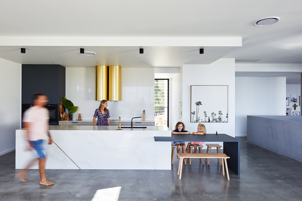 This is an example of a modern galley open plan kitchen in Gold Coast - Tweed with an undermount sink, flat-panel cabinets, light wood cabinets, white splashback, black appliances, concrete floors, with island, grey floor and white benchtop.