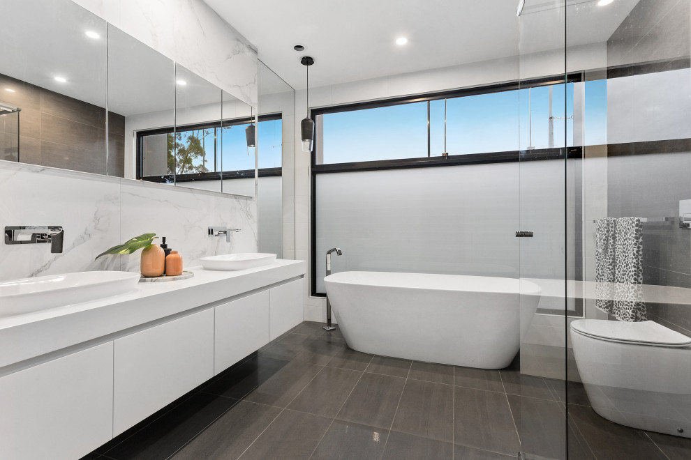 Photo of a large contemporary master bathroom in Melbourne with white cabinets, a freestanding tub, a corner shower, white tile, porcelain floors, marble benchtops, grey floor, a hinged shower door, white benchtops, flat-panel cabinets, a one-piece toilet and a vessel sink.