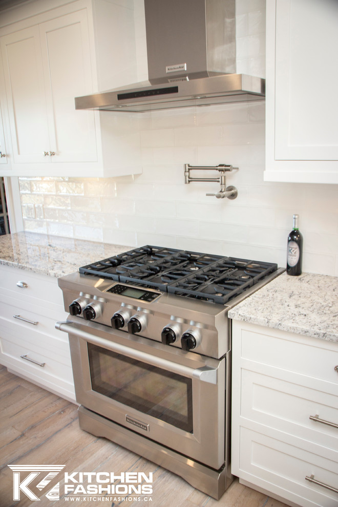 Large transitional u-shaped light wood floor eat-in kitchen photo with a farmhouse sink, shaker cabinets, white cabinets, granite countertops, white backsplash, subway tile backsplash, stainless steel appliances and no island