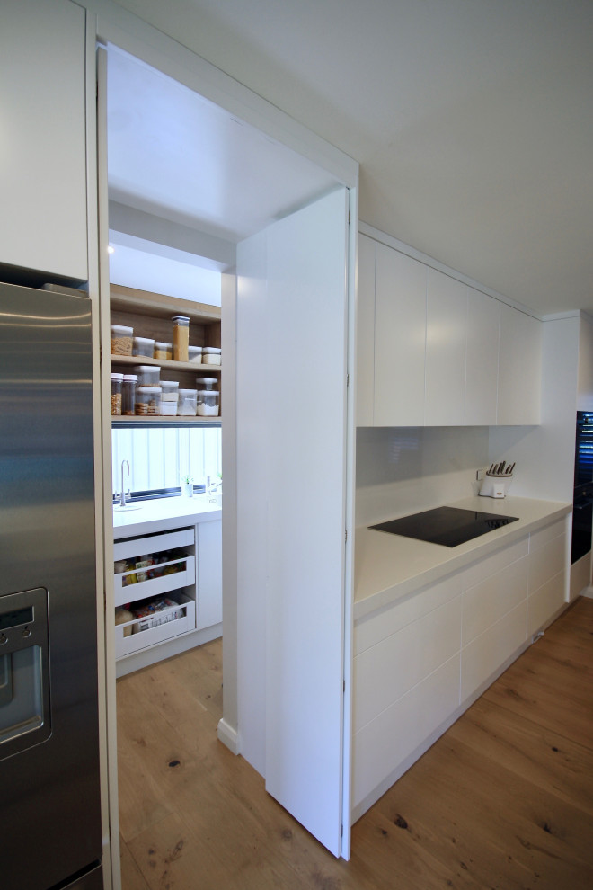 Design ideas for a large modern single-wall open plan kitchen in Sydney with a drop-in sink, flat-panel cabinets, white cabinets, quartz benchtops, white splashback, marble splashback, black appliances, medium hardwood floors, with island, brown floor and white benchtop.
