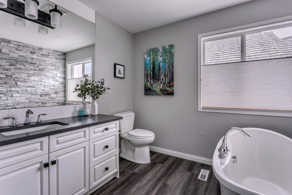 Mid-sized transitional master bathroom in Calgary with shaker cabinets, white cabinets, a freestanding tub, a two-piece toilet, white tile, ceramic tile, grey walls, vinyl floors, an undermount sink, engineered quartz benchtops and grey floor.