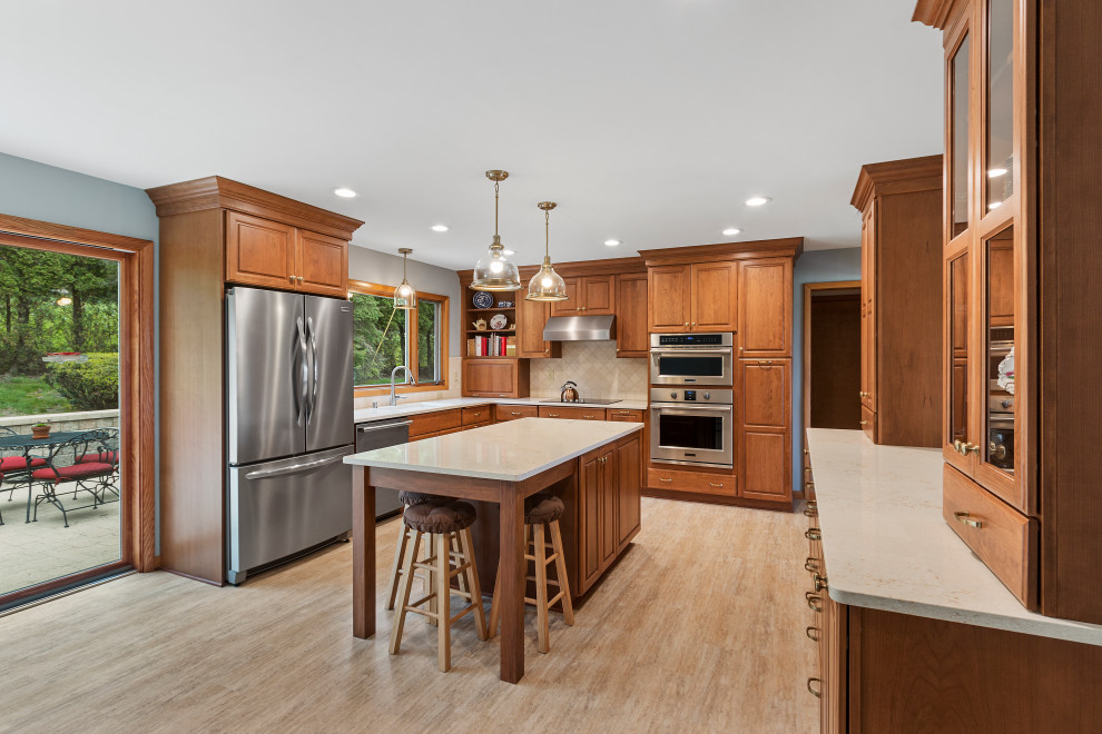 Photo of a mid-sized traditional l-shaped eat-in kitchen in Milwaukee with an undermount sink, raised-panel cabinets, brown cabinets, quartz benchtops, beige splashback, stone tile splashback, stainless steel appliances, vinyl floors, with island, brown floor and beige benchtop.