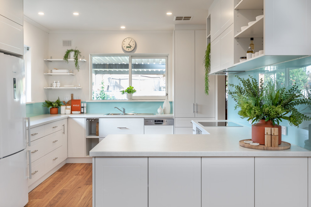 This is an example of a contemporary u-shaped kitchen in Adelaide with a drop-in sink, flat-panel cabinets, white cabinets, panelled appliances, medium hardwood floors, a peninsula, brown floor and white benchtop.