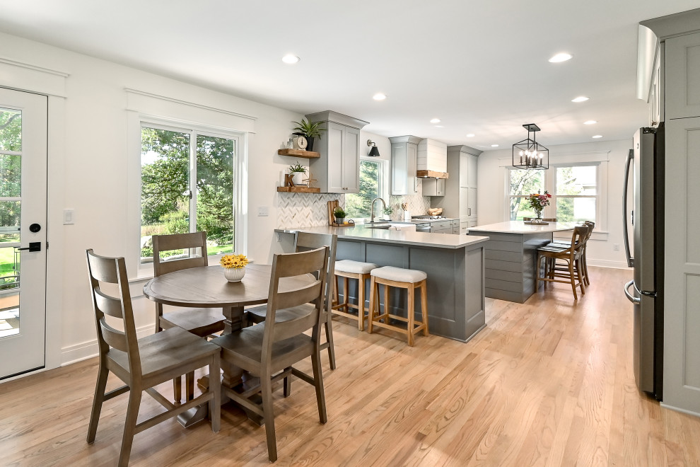 This is an example of an expansive country l-shaped eat-in kitchen in Milwaukee with a farmhouse sink, shaker cabinets, grey cabinets, quartz benchtops, white splashback, marble splashback, stainless steel appliances, medium hardwood floors, with island, brown floor and grey benchtop.