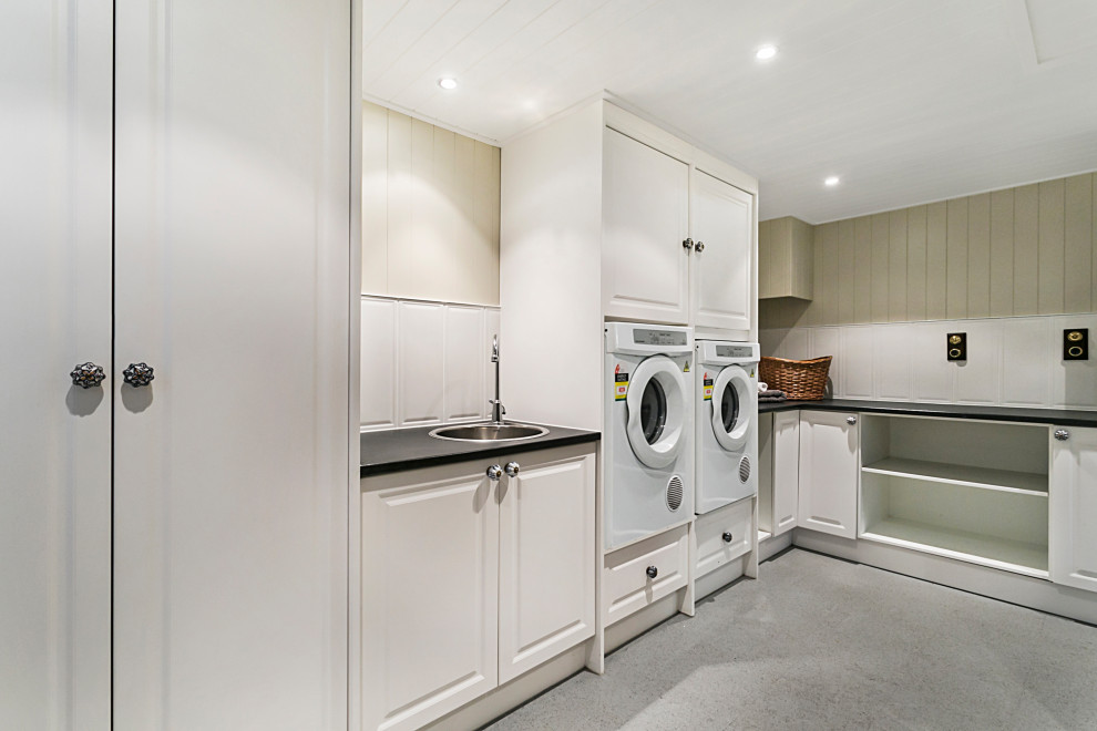 This is an example of an expansive traditional l-shaped dedicated laundry room in Brisbane with a drop-in sink, shaker cabinets, white cabinets, laminate benchtops, white splashback, ceramic splashback, beige walls, vinyl floors, a side-by-side washer and dryer, grey floor, black benchtop, timber and planked wall panelling.