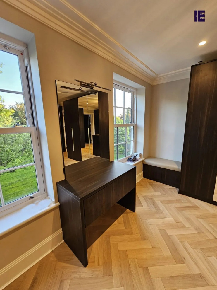 Photo of a large modern walk-in wardrobe in London with glass-front cabinets, dark wood cabinets and plywood floors.