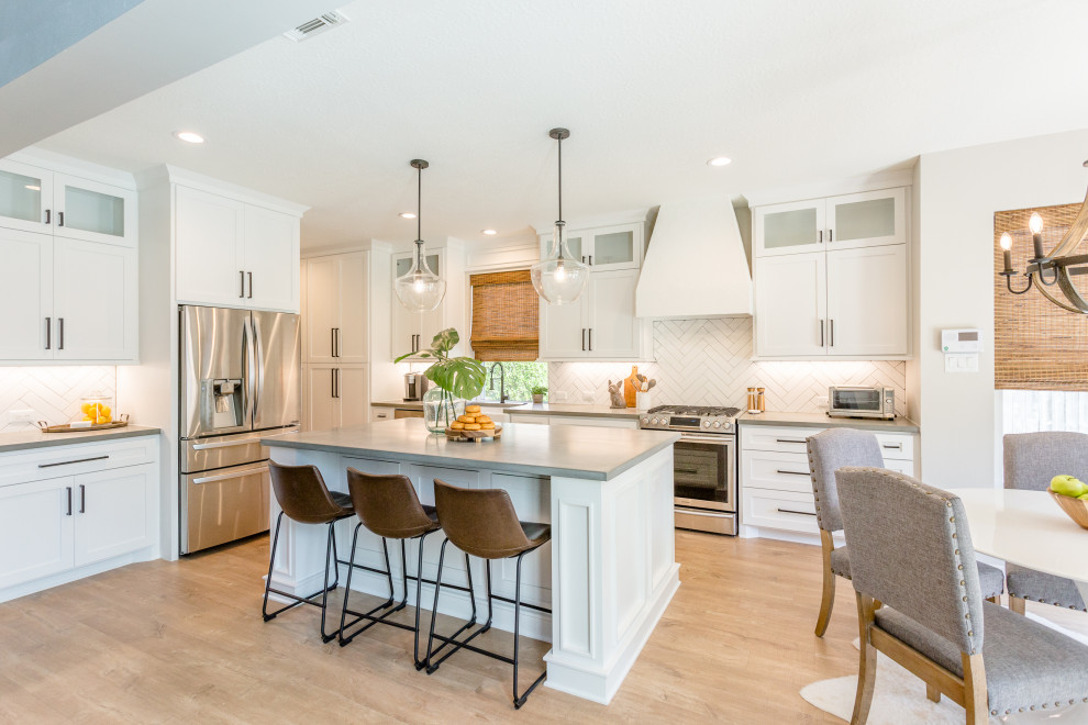 This is an example of a large transitional l-shaped eat-in kitchen in Houston with a farmhouse sink, shaker cabinets, white cabinets, white splashback, subway tile splashback, stainless steel appliances, light hardwood floors, with island, beige floor and grey benchtop.
