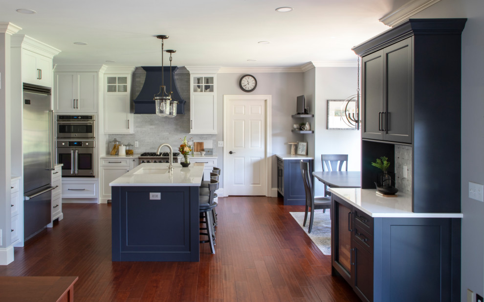 Example of a transitional medium tone wood floor kitchen design with an undermount sink, shaker cabinets, quartz countertops, gray backsplash, porcelain backsplash, stainless steel appliances, an island and white countertops