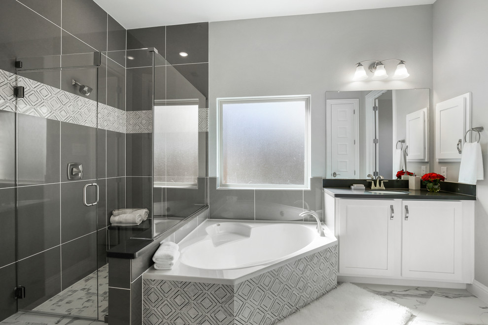 Mid-sized traditional master bathroom in Dallas with recessed-panel cabinets, white cabinets, a drop-in tub, a corner shower, gray tile, ceramic tile, grey walls, ceramic floors, an undermount sink, solid surface benchtops, white floor, a hinged shower door and black benchtops.