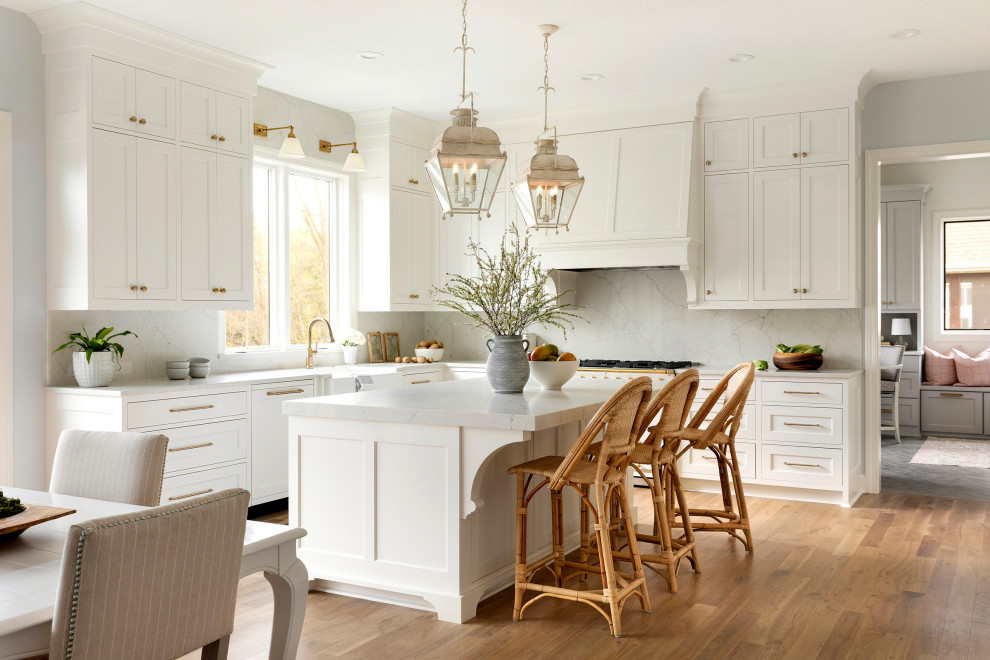 This is an example of an expansive traditional u-shaped eat-in kitchen in Minneapolis with a farmhouse sink, recessed-panel cabinets, white cabinets, quartz benchtops, white splashback, engineered quartz splashback, white appliances, light hardwood floors, with island, brown floor and white benchtop.