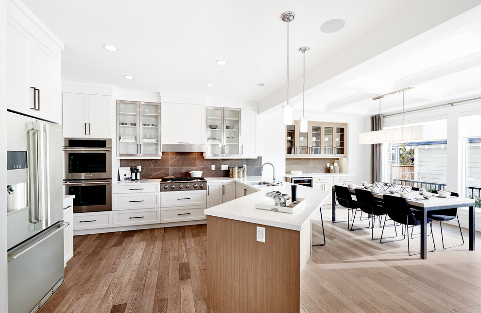 This is an example of a large transitional u-shaped eat-in kitchen in Vancouver with a double-bowl sink, white cabinets, granite benchtops, brown splashback, ceramic splashback, stainless steel appliances, light hardwood floors, a peninsula and shaker cabinets.