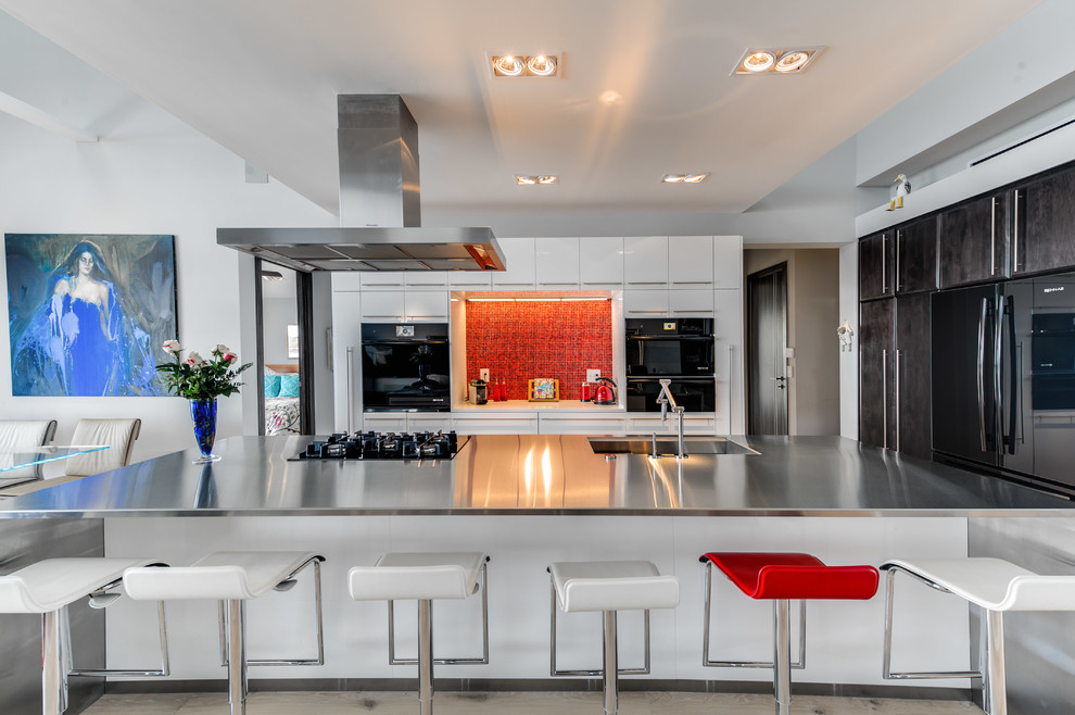 Large contemporary galley open plan kitchen in Other with an integrated sink, flat-panel cabinets, white cabinets, stainless steel benchtops, red splashback, glass tile splashback, black appliances, light hardwood floors and with island.