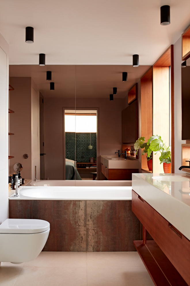 This is an example of a medium sized retro ensuite bathroom in Sussex with flat-panel cabinets, medium wood cabinets, a built-in bath, a wall mounted toilet, mirror tiles, limestone flooring, a built-in sink, solid surface worktops, white worktops, a feature wall, double sinks and a floating vanity unit.
