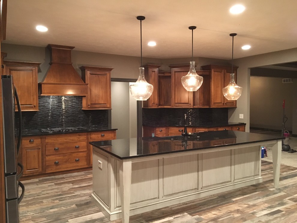 Example of a large transitional l-shaped porcelain tile eat-in kitchen design in Omaha with an undermount sink, raised-panel cabinets, medium tone wood cabinets, granite countertops, black backsplash, matchstick tile backsplash, stainless steel appliances and an island