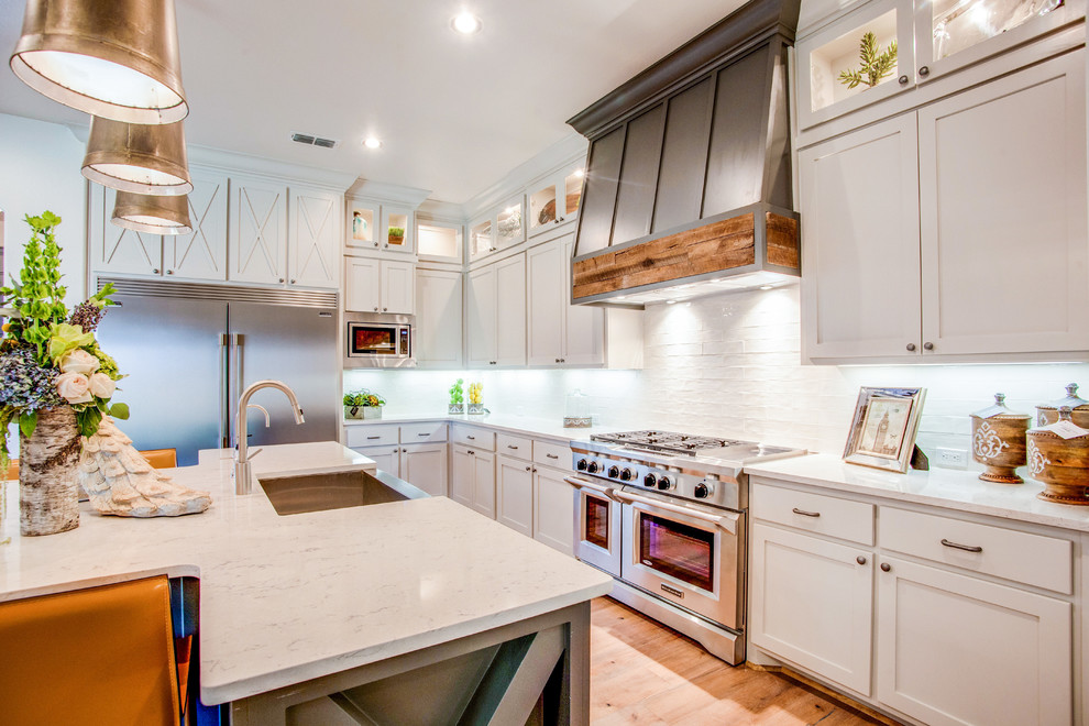 Country u-shaped kitchen in Austin with a farmhouse sink, shaker cabinets, white cabinets, white splashback, stainless steel appliances, light hardwood floors and with island.