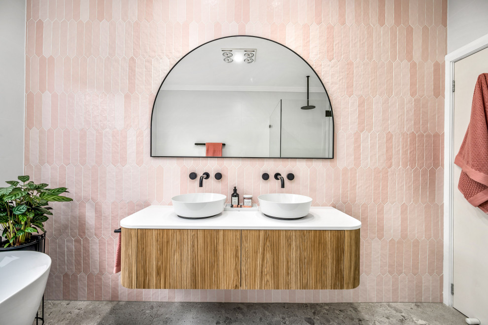 Photo of a mid-sized beach style master bathroom in Central Coast with furniture-like cabinets, medium wood cabinets, a freestanding tub, a corner shower, a one-piece toilet, pink tile, white walls, a vessel sink, grey floor, a hinged shower door, white benchtops, a niche, a double vanity and a floating vanity.