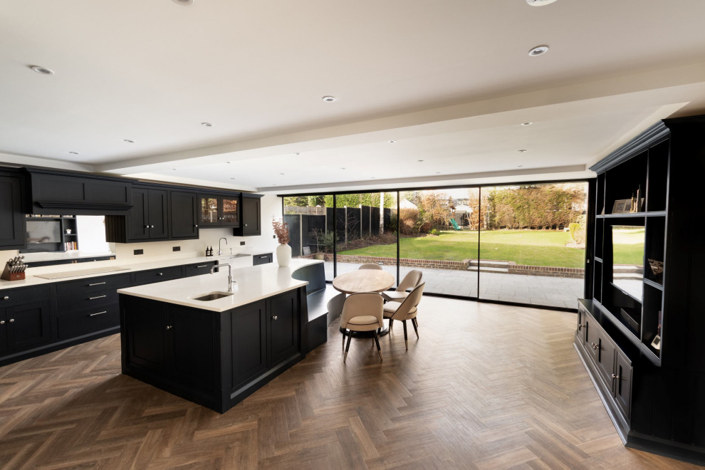 Photo of an expansive contemporary l-shaped open plan kitchen in Hertfordshire with a farmhouse sink, shaker cabinets, black cabinets, quartzite benchtops, metallic splashback, glass sheet splashback, black appliances, medium hardwood floors, with island, brown floor and white benchtop.