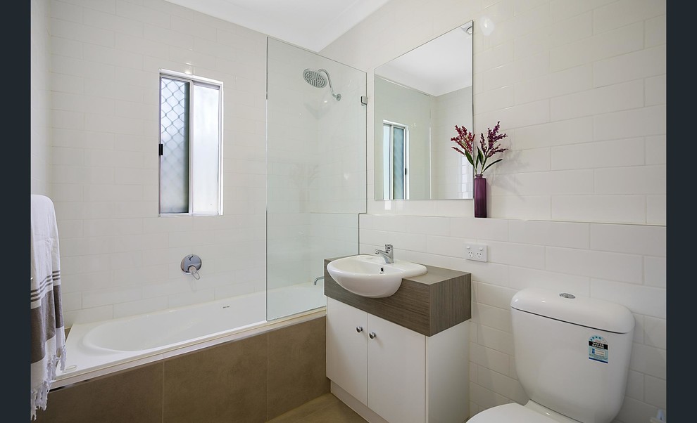Mid-sized contemporary kids bathroom in Gold Coast - Tweed with white cabinets, a drop-in tub, a shower/bathtub combo, a one-piece toilet, white tile, subway tile, white walls, ceramic floors, a drop-in sink, laminate benchtops and brown floor.