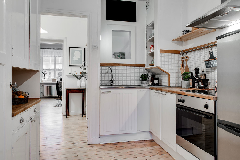 Inspiration for a mid-sized scandinavian l-shaped separate kitchen in Gothenburg with wood benchtops, stainless steel appliances, light hardwood floors, no island, white cabinets, white splashback, mosaic tile splashback and a double-bowl sink.