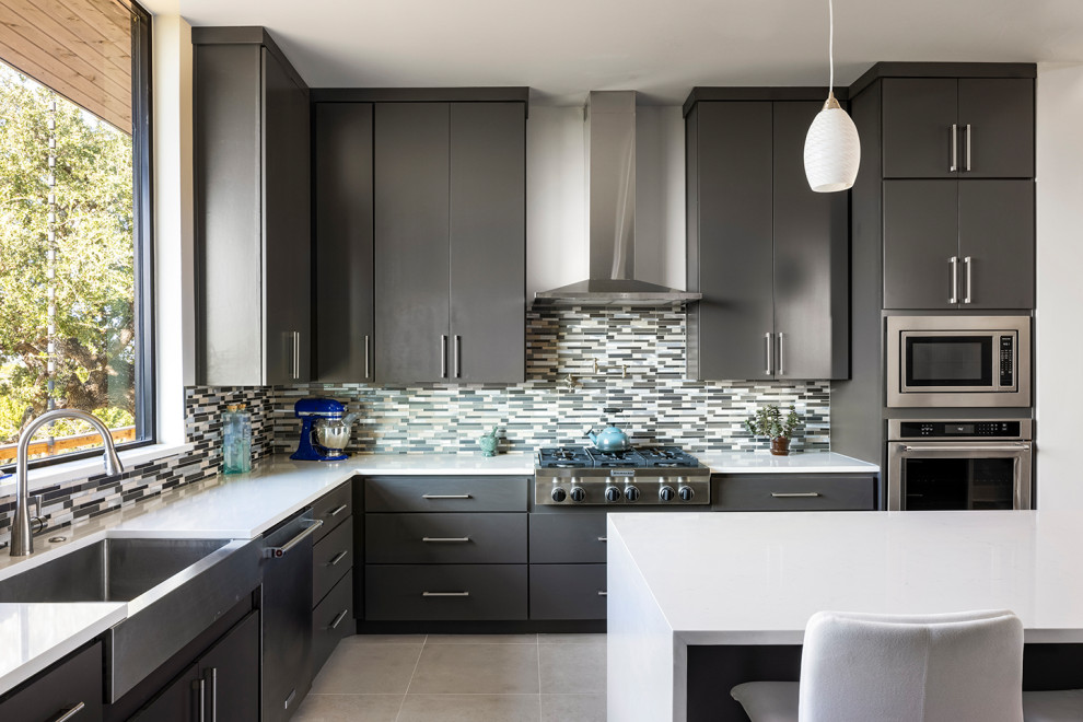 Contemporary l-shaped kitchen in Austin with a belfast sink, flat-panel cabinets, black cabinets, multi-coloured splashback, matchstick tiled splashback, stainless steel appliances, an island, grey floors and white worktops.