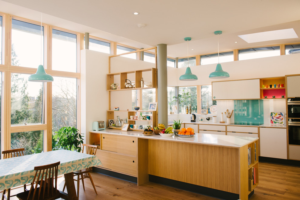 Large midcentury u-shaped open plan kitchen in Manchester with flat-panel cabinets, white cabinets, solid surface benchtops, blue splashback, glass sheet splashback, stainless steel appliances, no island and white benchtop.
