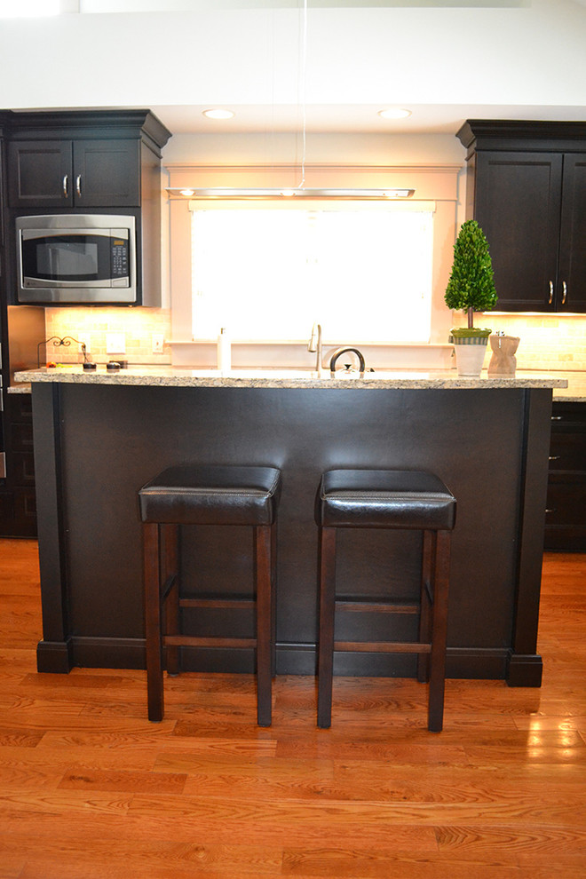 Example of a large transitional galley medium tone wood floor enclosed kitchen design in Columbus with an undermount sink, recessed-panel cabinets, dark wood cabinets, granite countertops, beige backsplash, stone tile backsplash, stainless steel appliances and an island