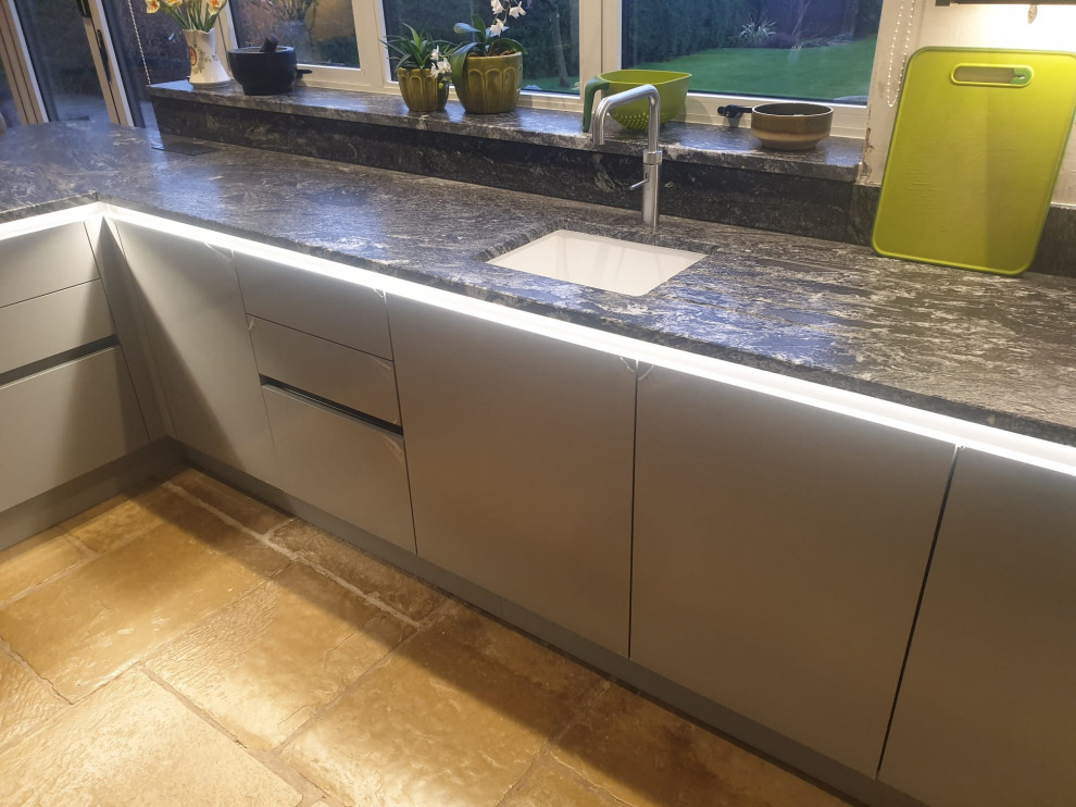 This is an example of a medium sized contemporary grey and black l-shaped kitchen/diner in West Midlands with flat-panel cabinets, granite worktops, black splashback, engineered quartz splashback, stainless steel appliances, black worktops, feature lighting, a built-in sink, black cabinets, travertine flooring, a breakfast bar, beige floors and a vaulted ceiling.