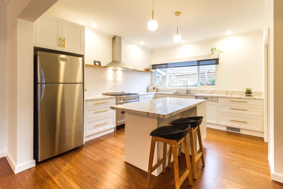 Mid-sized transitional u-shaped kitchen pantry in Melbourne with a drop-in sink, shaker cabinets, white cabinets, quartz benchtops, white splashback, ceramic splashback, stainless steel appliances, medium hardwood floors, with island and brown floor.