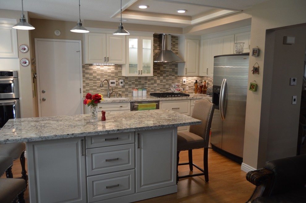 Photo of a small traditional l-shaped open plan kitchen in Orange County with a double-bowl sink, raised-panel cabinets, white cabinets, granite benchtops, beige splashback, glass tile splashback, stainless steel appliances, light hardwood floors, with island, beige floor and grey benchtop.