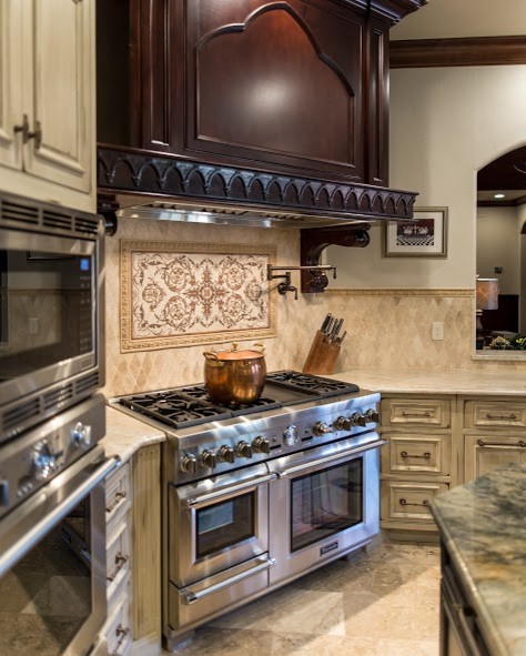 Photo of a large u-shaped eat-in kitchen in Oklahoma City with a farmhouse sink, beaded inset cabinets, beige cabinets, granite benchtops, beige splashback, travertine splashback, stainless steel appliances, travertine floors, with island and beige floor.