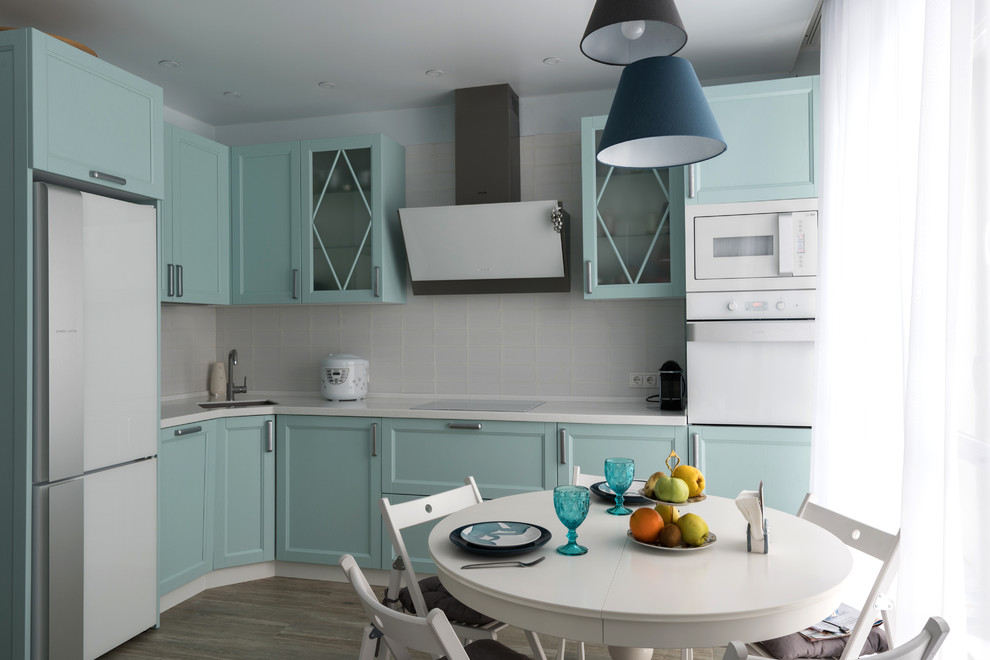This is an example of a mid-sized transitional l-shaped separate kitchen in Moscow with an integrated sink, recessed-panel cabinets, turquoise cabinets, solid surface benchtops, white splashback, subway tile splashback, white appliances, laminate floors, no island, brown floor and white benchtop.