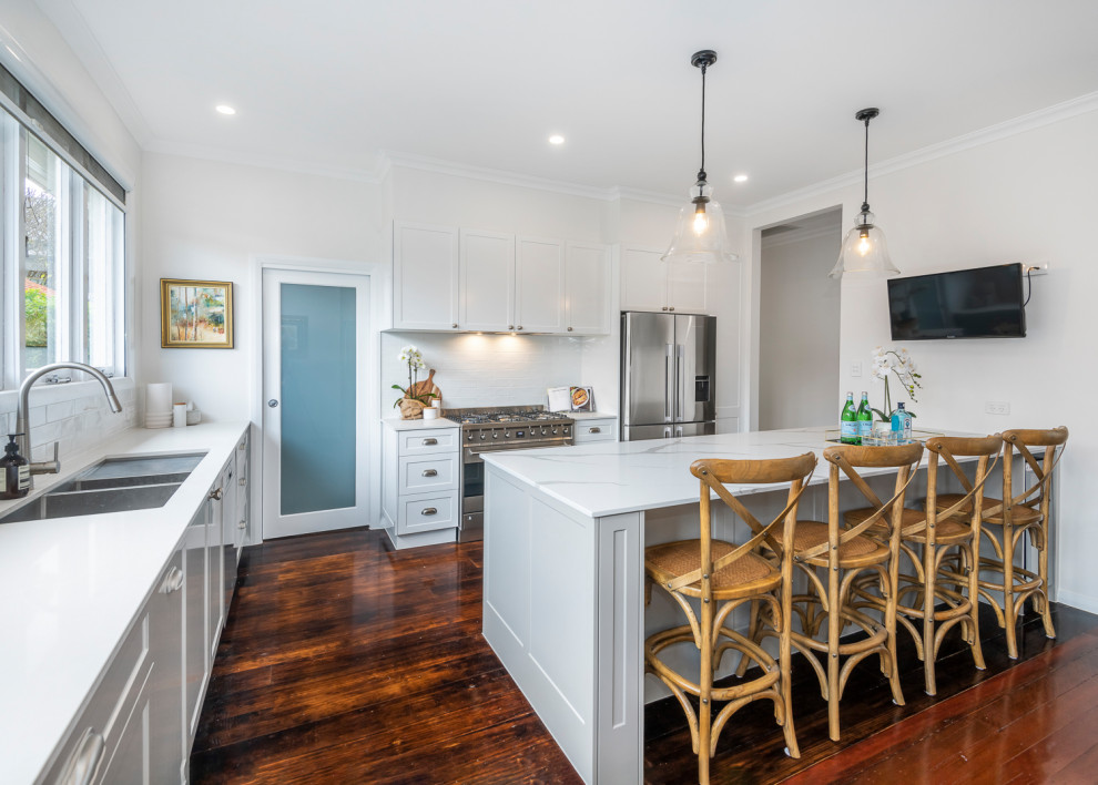 This is an example of a large transitional l-shaped open plan kitchen in Brisbane with a double-bowl sink, shaker cabinets, grey cabinets, solid surface benchtops, white splashback, subway tile splashback, stainless steel appliances, dark hardwood floors, with island and white benchtop.