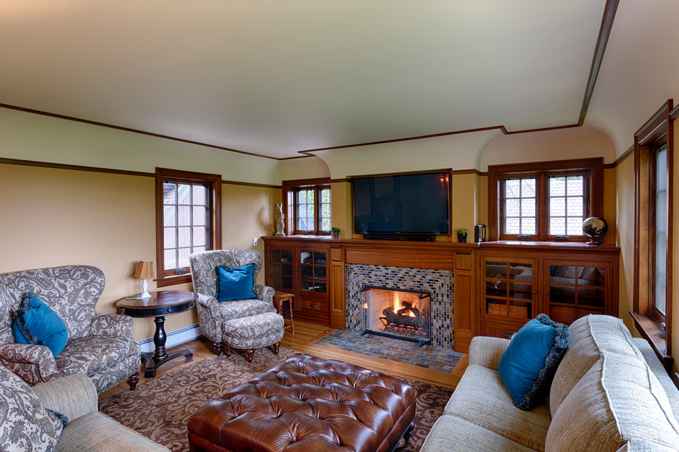 Inspiration for a mid-sized traditional enclosed family room in Seattle with yellow walls, light hardwood floors, a standard fireplace, a tile fireplace surround, a wall-mounted tv and beige floor.