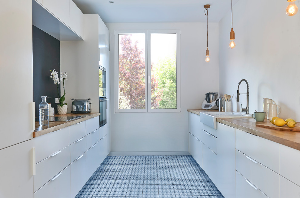 Photo of a mid-sized scandinavian galley separate kitchen in Paris with a farmhouse sink, flat-panel cabinets, white cabinets, black splashback, white appliances, no island, blue floor and beige benchtop.